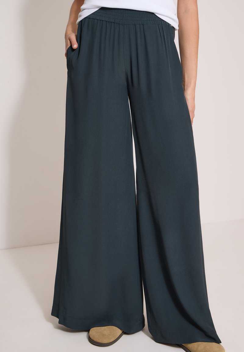Personne portant un pantalon large sombre avec taille élastique, un haut blanc et des chaussures marron clair, debout avec une main dans la poche.