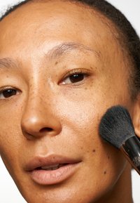 Nærbilde av et ansikt med varm hudtone og fregner, som bruker en sminkepute med svarte børster på kinnet. Glatt tekstur, naturlig utseende.