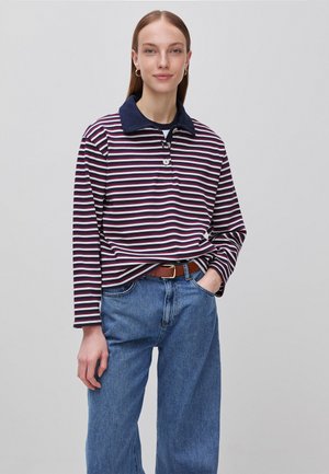 Młoda kobieta z prostymi włosami, ubrana w pasiastą koszulkę polo z długim rękawem, niebieskie jeansy i brązowy pasek, stoi z jedną ręką w kieszeni.