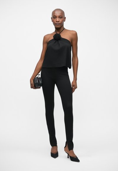 Zwarte haltertop met een bloemaccent, gecombineerd met strakke zwarte leggings en zwarte hoge hakken. De outfit heeft een gladde textuur en een slanke silhoeet.