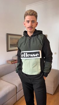 Grön och svart färgblockerad jacka med huva, frontdragkedja och stor vit "ellesse"-logga. Svarta byxor kompletterar outfitens design.
