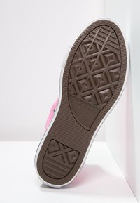 Růžové plátěné tenisky s bílou gumovou podrážkou, které mají texturovanou hnědou podrážku se geometrickými vzory a detail loga.