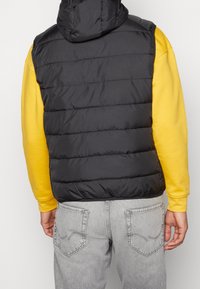 Svart pufferväst med horisontell sömnad, dragkedja fram och huva, bärs över en gul sweatshirt, i kombination med ljusgrå jeans.