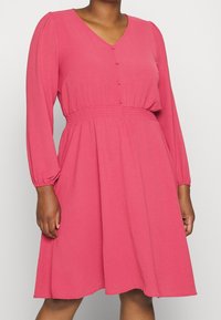 Rosa klänning med långa ärmar och V-ringning, knapppdetalj och rynkad midja. Tillverkad av mjukt material med en avslappnad A-linjeform.
