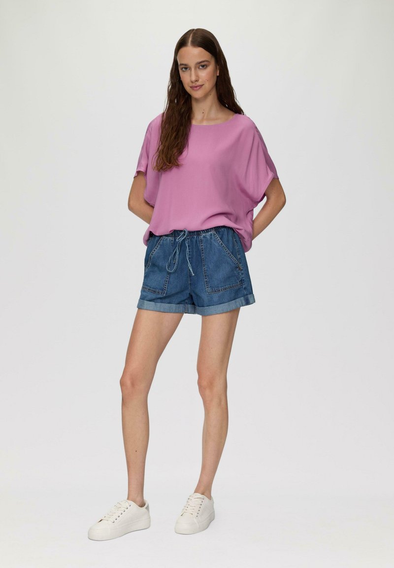 Roze loszittend shirt met een brede halslijn, gecombineerd met blauwe denimshorts met opgerolde mouwen en een trekkoord in de taille. Witte sneakers.