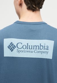 Οπίσθια όψη ατόμου που φορά μπλε πουκάμισο με ελαφρώς γαλάζιο ορθογώνιο λογότυπο που γράφει «Columbia Sportswear Company» στο πάνω μέρος της πλάτης.