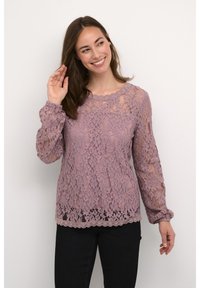 Spitzenoberteil in sanftem Lila mit langen Ärmeln, gewelltem Ausschnitt und floralem Muster. Kombiniert mit schwarzen Hosen; durchscheinende Textur sichtbar.