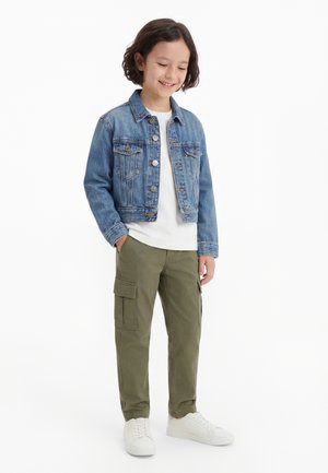 Kind lächelt, steht mit Händen in den Taschen der olivgrünen Cargohose, trägt eine blaue Jeansjacke, ein weißes Hemd und weiße Turnschuhe.