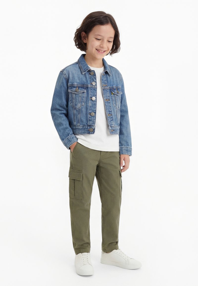 Copil zâmbind, stând cu mâinile în buzunarele pantalonilor cargo verde măsliniu, purtând o jachetă albastră din denim, cămașă albă și adidași albi.