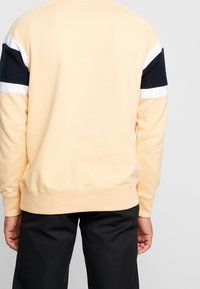 Person som bär en beige tröja med marinblå och vita ränder på ärmarna, i kombination med svarta byxor, visad bakifrån.