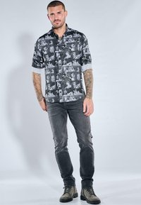 Schwarzes und weißes gemustertes Hemd mit verschiedenen künstlerischen Motiven, kombiniert mit grauen Jeans und grünen Stiefeln. Glatter Stoff, Slim-Fit-Design.
