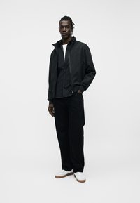 Νεαρός άνδρας που στέκεται με το χέρι στην τσέπη, φοράει μαύρο σακάκι, μαύρο πουκάμισο, μαύρο παντελόνι και λευκά αθλητικά παπούτσια με καφέ σόλες.