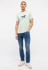 Jasnozielona bawełniana koszulka z graficznym motywem konia w kolorze granatowym. Nosi ją z niebieskimi dżinsami i białymi trampkami z beżowymi akcentami oraz teksturowaną podeszwą.