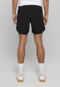 Czarne sportowe spodenki z elastycznym pasem, lekkim materiałem i subtelną naszywką z logo z tyłu. Noszone z białymi skarpetami i butami.