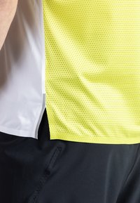 Lekka koszulka biegowa z żółtej siateczki z małymi, teksturowanymi wzorami, połączona z gładką białą boczną wstawką.