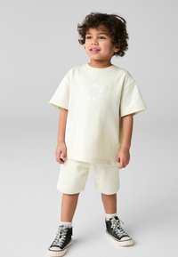 Lys cremefarget t-shirt med korte ermer og cirkulært grafisk design, parret med matchende shorts og sorte høj-top sneakers med hvid sål.