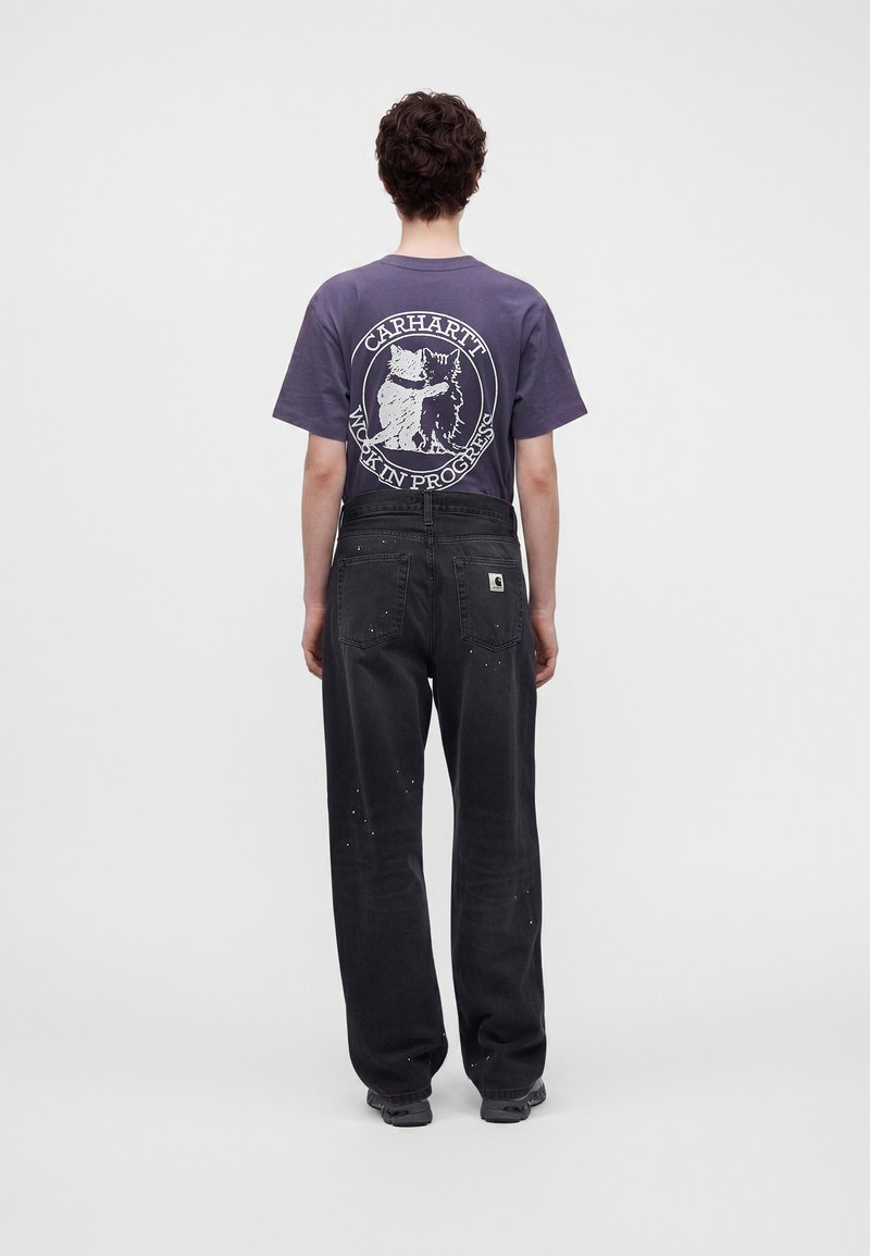 Persona con capelli corti e scuri che indossa una maglietta Carhartt viola con grafica di gatto e pantaloni neri a gamba larga con macchie di vernice, di spalle.