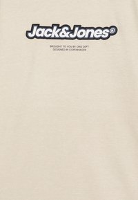 T-shirt in cotone beige con un logo nero prominente "Jack & Jones" e un simbolo di marchio circolare sopra, con testo più piccolo sotto.
