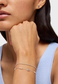 Pulseras de plata con acentos de cuentas en una muñeca; una de ellas presenta un pequeño encanto de pez. El modelo tiene la piel suave y lleva una blusa azul claro.