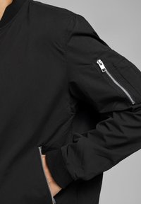 Černá bomber bunda s hladkým materiálem, žebrovanými manžetami, bočními kapsami a zipovou kapsou na rukávu se stříbrným kováním.
