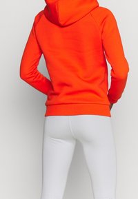 Sweatshirt orange vif à capuche, manches longues et ourlet côtelé, associé à des leggings blancs ajustés avec des détails perforés.