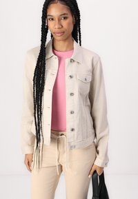 Beige denim jacket with button closures, two chest pockets, and subtle texture, worn over a pink shirt and paired with beige pants.