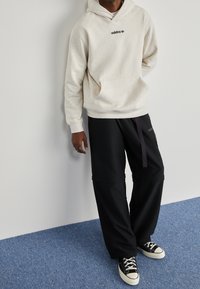 Beige Kapuzenpullover mit schwarzem "adidas"-Logo, frontale Tasche; kombiniert mit schwarzen weit geschnittenen Hosen, lila Kordelzug und schwarzen Leinenschuhen.