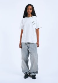 Bílý tričko s krátkým rukávem a grafickými detaily, spárované s volnými světle modrými džíny. Model má na sobě černé boty se zdobnými prvky.