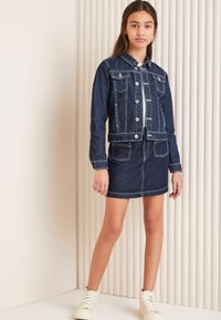 Temno modra jeans jakna z belimi šivi in dvema žepoma na prsih, v kombinaciji z ujemajočo se jeans krilom. Model nosi bež visoke superge.