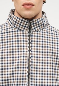 Κοντινό πλάνο ατόμου που φοράει καφέ και μπλε καρό μπουφάν με μαύρο φερμουάρ που γράφει "WEEKEND", ζιπ κουστούμι μέχρι τον λαιμό.