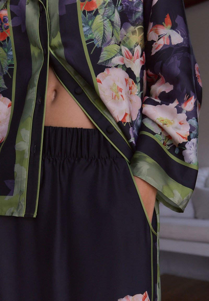 Blusa in seta floreale con base nera, dettagli verdi e chiusura con bottoni. Abbinata a una gonna nera con vita elastica, che mostra una texture liscia.