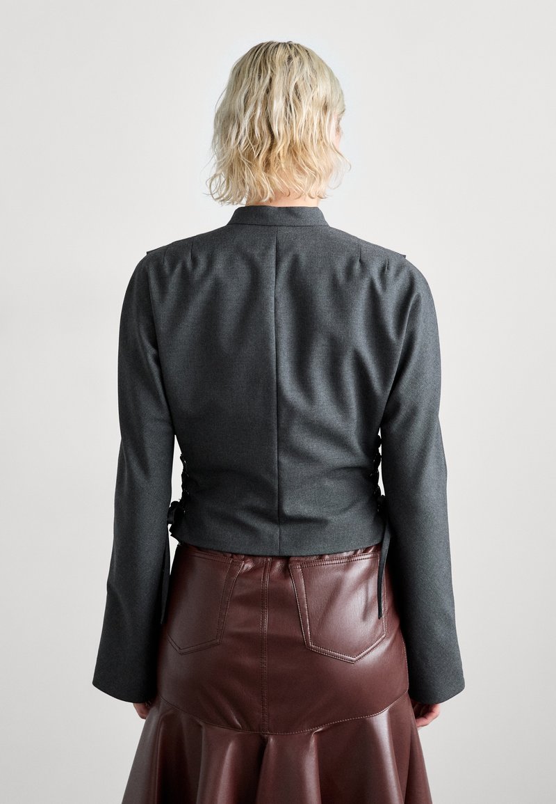 Giacca tailored grigia scura con maniche lunghe, caratterizzata da un colletto alto e lacci sul retro; abbinata a una gonna in ecopelle bordeaux con tasche.