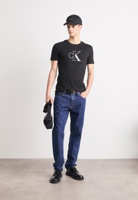 Must musta särk valge "CK" logoga, sinised teksad, must vöö, mustad kingad ja must müts, hoides vasakus käes peakomplekti.