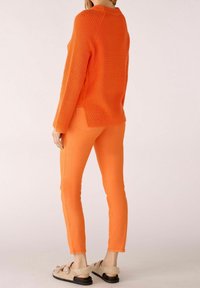 Femme portant un pull en tricot orange vif et un pantalon slim assorti, debout avec le dos partiellement tourné, des sandales beiges en fourrure aux pieds.