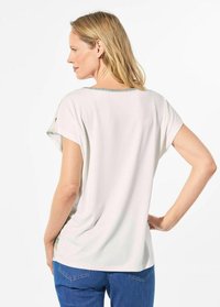 Witte blouse met korte mouwen en een ronde halslijn, met contrasterende stiksels langs de halslijn en een gladde textuur. Gecombineerd met blauwe jeans.