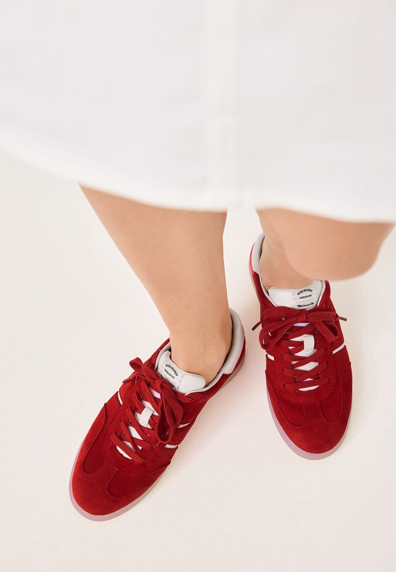 Personne portant des baskets en daim rouge avec des accents blancs et des lacets, debout sur une surface de couleur claire, visible des genoux vers le bas.