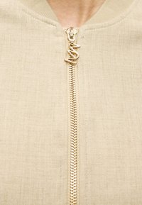 Η μπεζ μπέρμπον μπουφάν διαθέτει ριμπ ύφασμα στον γιακά και χρυσό φερμουάρ με στυλιζαρισμένο τράβηγμα. Ομαλή υφή με διακριτικό μοτίβο.