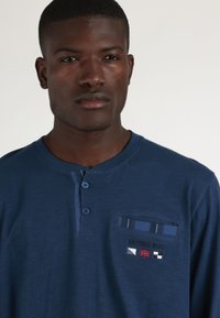 Camisa de manga larga azul marino con diseño tipo henley, dos botones en el cuello y un bolsillo que presenta un logotipo bordado y acentos a rayas.