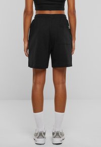 Personne portant un short noir à taille élastique, des chaussettes blanches montantes et des baskets grises et blanches, debout devant un fond gris uni.