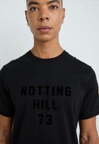 Svart bomull t-skjorte med en hevet, fløyelsaktig trykk av ordene "NOTTING HILL 73" i fete bokstaver over brystet.