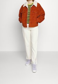 Kordové sako v rezavě oranžové barvě se bílým sherpa límcem, pruhované zeleno-černé tričko, off-white džíny se třepenými okraji a levandulové tenisky.