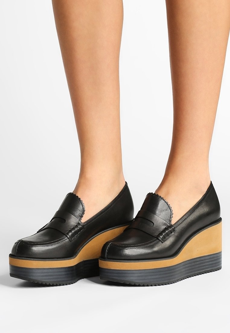 Mocassins à plateforme en cuir noir avec bords festonnés portés aux pieds, dotés de semelles compensées épaisses superposées camel et noir.