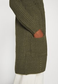 Personne portant un cardigan en maille côtelée vert olive avec la main dans la poche avant, associé à un pantalon blanc, visible de côté de la taille vers le bas.