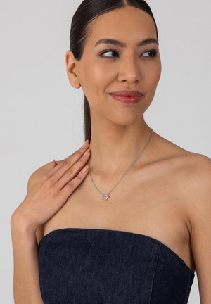 Junge Frau trägt ein trägerloses, dunkles Denim-Top und eine silberne Halskette mit Blumenanhänger, berührt die Halskette sanft und blickt mit einem leichten Lächeln nach rechts.