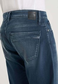 Jean en denim bleu foncé avec une texture lisse, doté d'un patch en cuir sur la ceinture et de deux poches arrière, avec une légère décoloration.