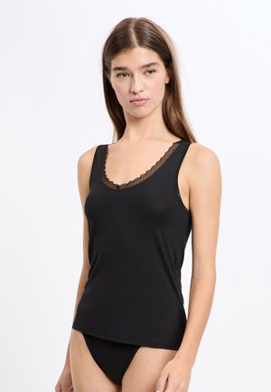 Młoda kobieta ubrana w czarny top bez rękawów z koronkowym wykończeniem i dopasowaną czarną bieliznę na jasnym, jednolitym tle.