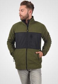 Fleece jacket in olive green with black paneling, zippered front, high collar, and chest pocket. Features two side pockets.
