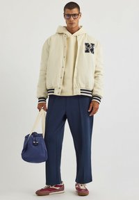 Kremowa kurtka bomberka z granatowymi paskami, beżowa bluza pod spodem, granatowe spodnie i bordowe sneakersy. Trzyma niebieską torbę z materiału.