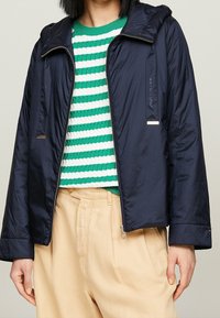 Personne portant une veste zippée bleu marine sur un pull rayé vert et blanc avec un pantalon large beige, se tenant devant un fond neutre.