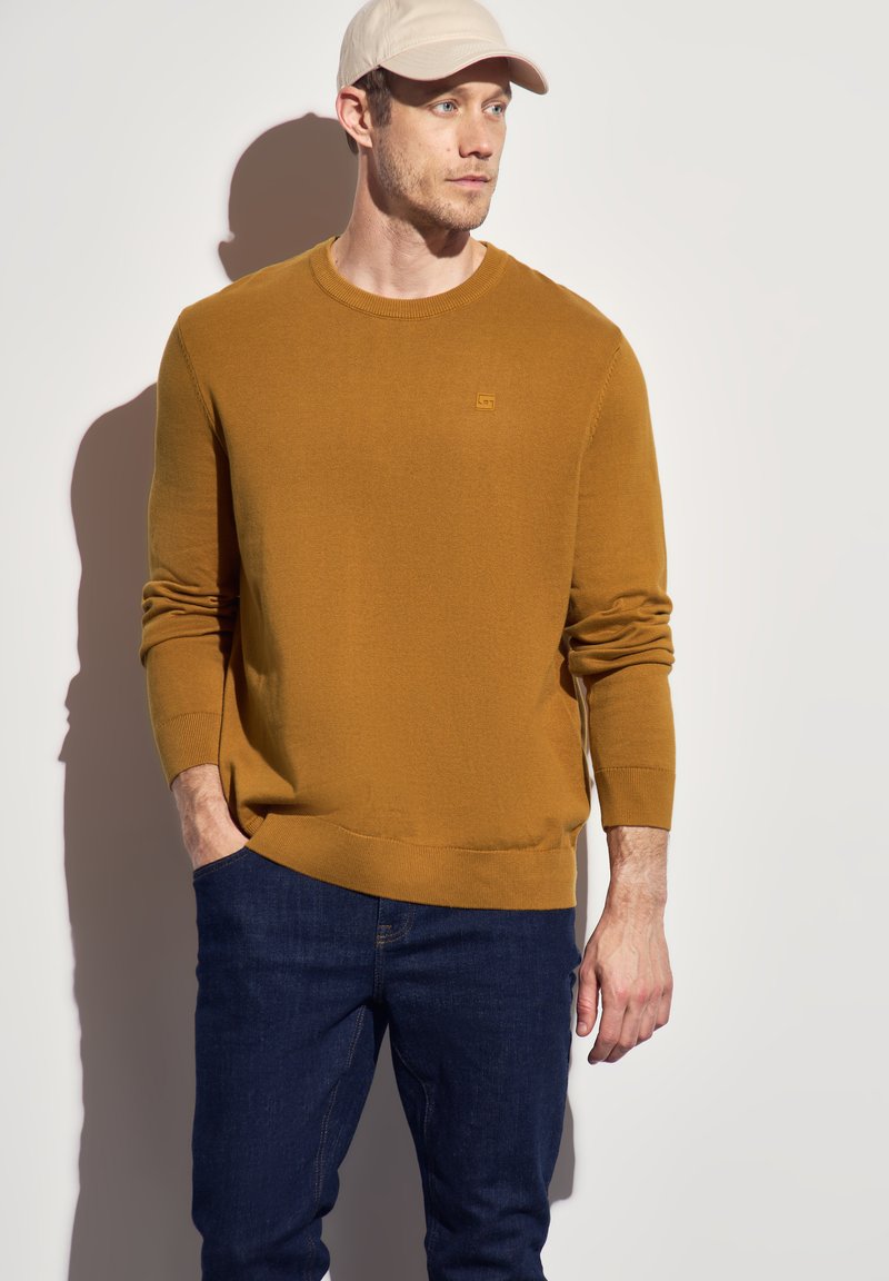 Senfgelber Strickpullover mit rundem Kragen und gerippten Bündchen, kombiniert mit dunkelblauen Jeans. Einfaches Logodesign auf der Brust.